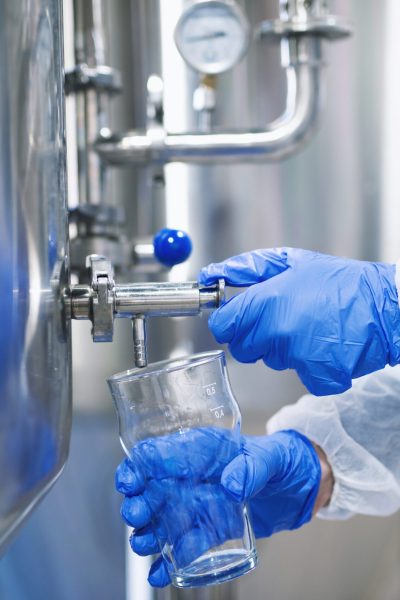 Close up view of hands in protective gloves opening tap valve and filling glass with samples from industrial machine reservoir.